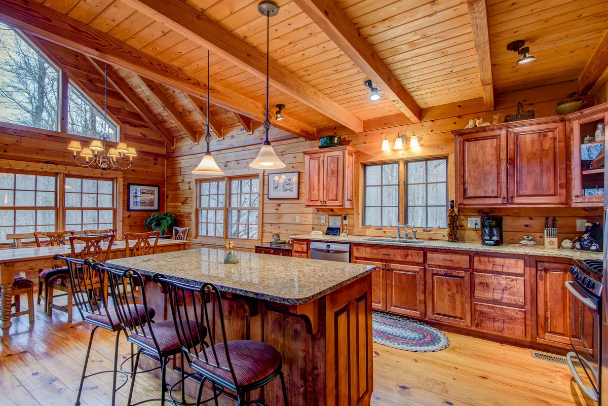 This image features a welcoming, open-concept kitchen and dining area, with a generous granite island, wooden cabinets, modern chandeliers, and large windows bathing the space in natural light. The adjacent dining area is visible, inviting gatherings and shared meals.
