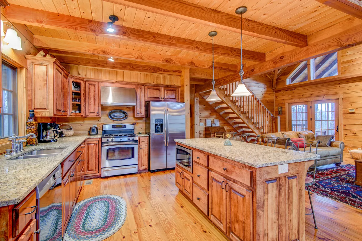 The image depicts a granite kitchen island surrounded by wooden cabinetry and stainless steel appliances, with a staircase and a cozy sitting area visible in the background. The kitchen is staged for meal prep and social engagement.