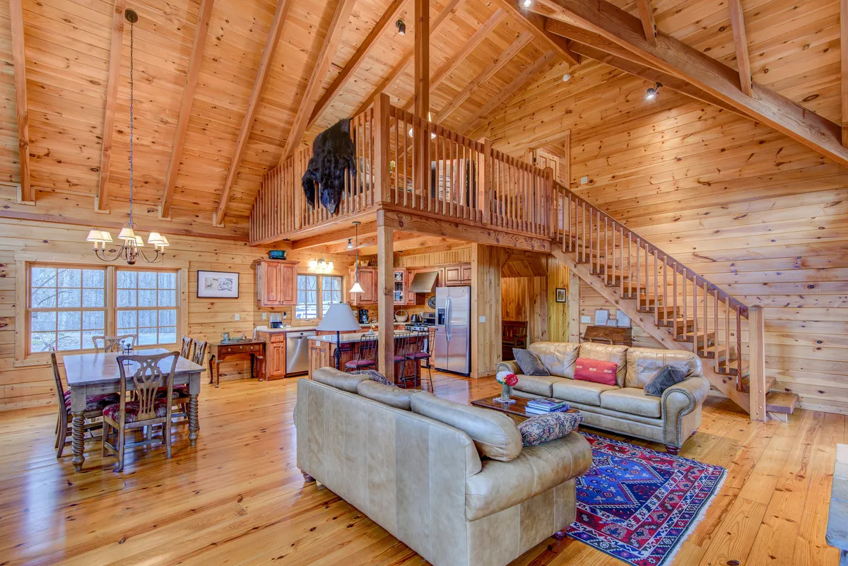 A spacious dining table sits beneath impressive wooden beams, with an open staircase and comfortable sofas nearby. The layout encourages gathering for meals or games, surrounded by a cozy, welcoming atmosphere.