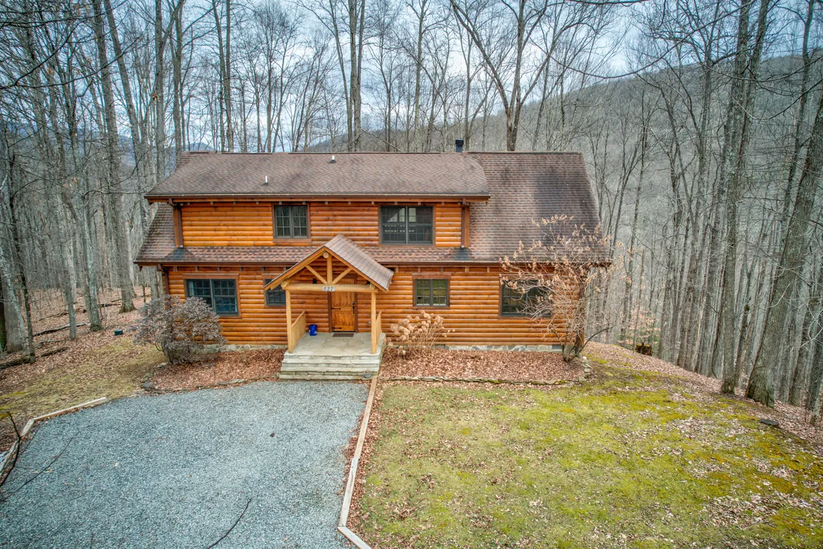 This image showcases the log cabin exterior nestled among mature trees, with a wide front porch and a gravel driveway in view. The setting suggests ample outdoor space for families to gather, kids to play, and easy access to nature trails right outside the door.