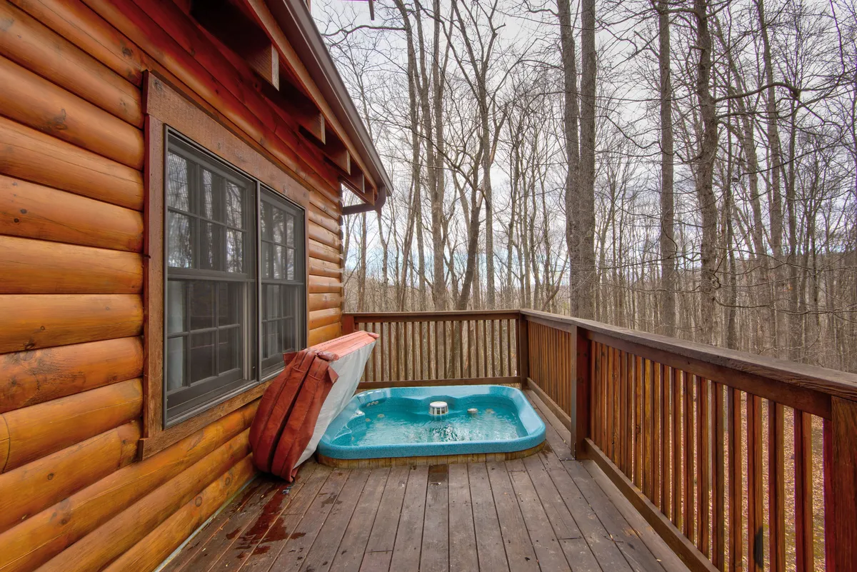 This image shows a serene outdoor hot tub nestled on a wooden deck attached to a log cabin, surrounded by tall trees. The view stretches into the forest, creating a secluded and peaceful atmosphere that's perfect for relaxation at any time of year.
