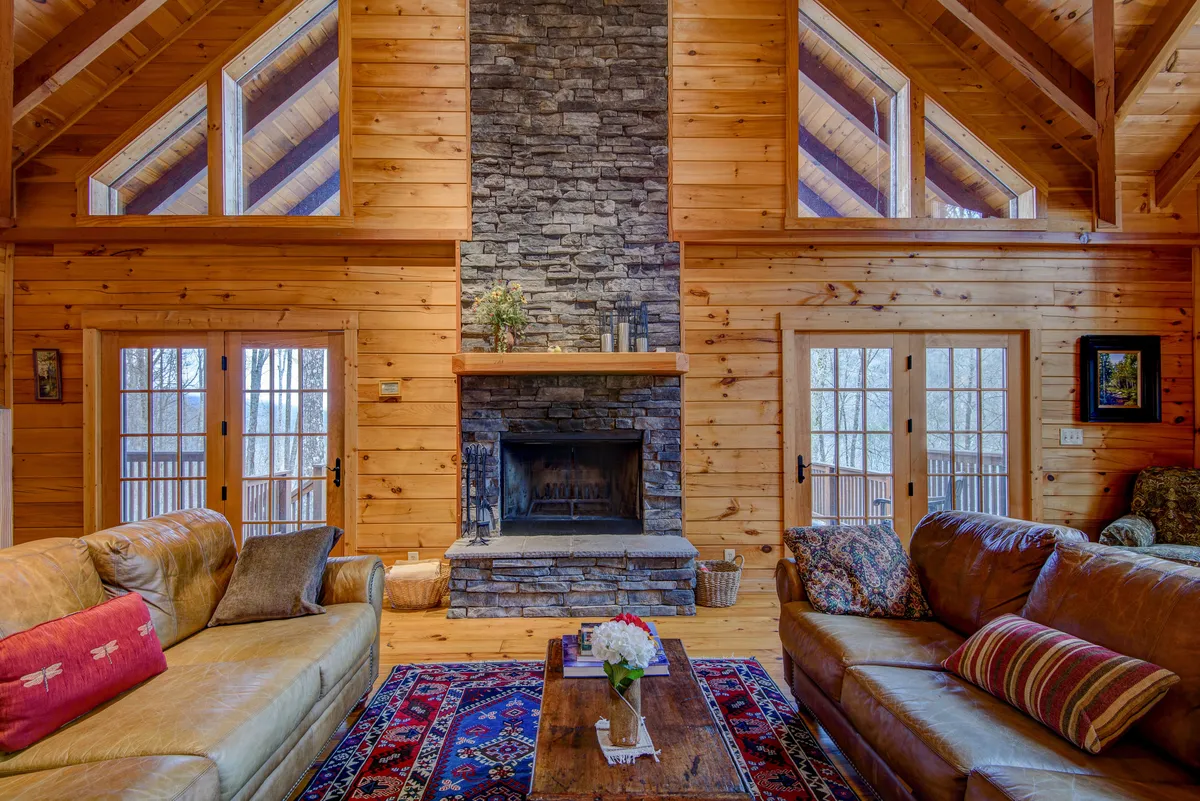 A beautiful living room featuring a grand stone fireplace set beneath soaring wooden beams. Plush sofas are arranged around a cozy rug, with large windows letting in natural light and offering glimpses of the surrounding estate. The warm wooden architecture and inviting seating make this an ideal setting for family gatherings and relaxing evenings.