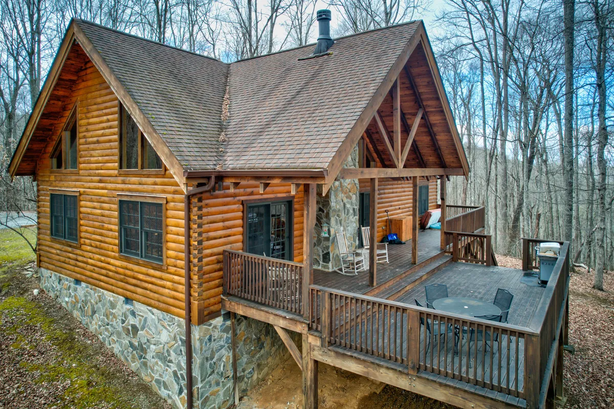 The hero image features a charming log cabin nestled among lush trees, with a spacious deck equipped with seating perfect for relaxing and taking in the mountain scenery. The sturdy stone foundation adds to the rustic appeal, while the natural surroundings hint at tranquil seclusion and opportunities for wildlife spotting. Soft sunlight bathes the deck, suggesting the perfect spot for enjoying coffee or gathering at sunset.