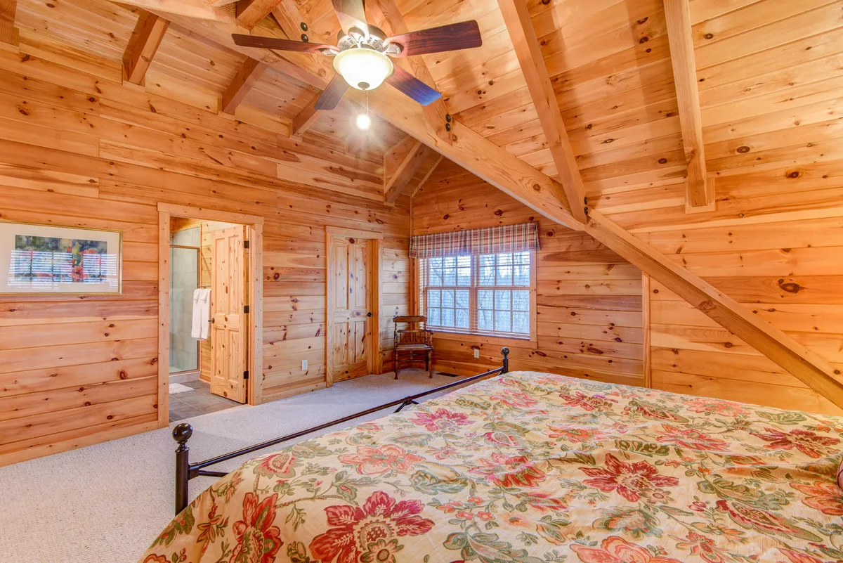 This bedroom features wooden ceiling beams, a floral-patterned bedspread, a window with checkered curtains, and a wooden chair. Natural light streams in through the window, creating a gentle, inviting ambiance that promises a serene start to the day.