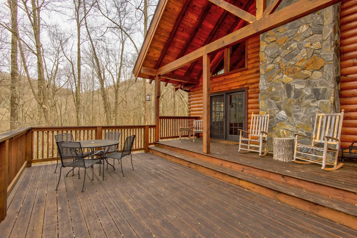 An exterior view of a wooden deck featuring rocking chairs, a stone chimney, and a dining table. The deck overlooks a scenic natural backdrop, inviting families to relax, enjoy meals outdoors, or gather together for stories and s'mores after a day of adventure.