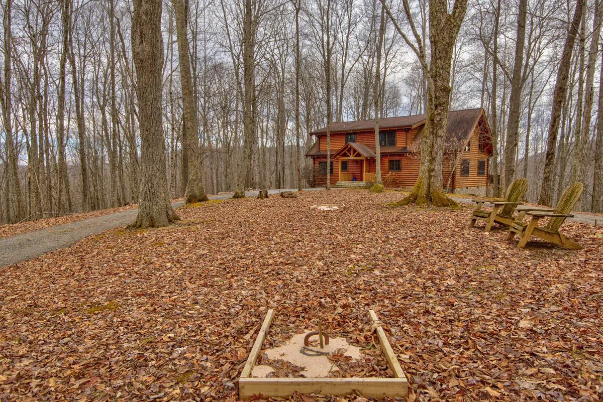 The image presents a log cabin framed by a leafy groundcover and surrounded by a dense tree canopy. To the side, an outdoor seating area encircles a rustic fire pit, inviting gatherings beneath the open sky.