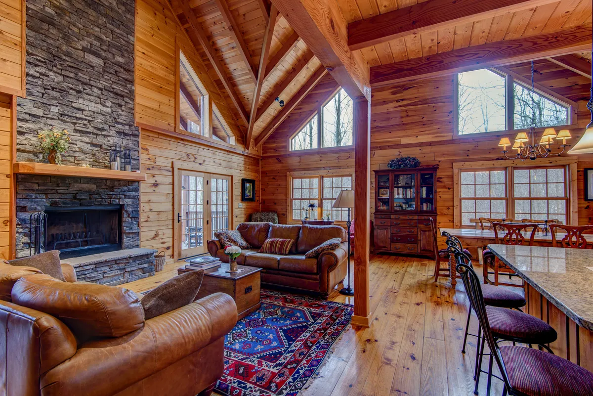 A spacious great room showcasing a large stone fireplace, sturdy wooden beams overhead, and abundant natural light through generous windows. Comfortable seating is arranged to invite conversation, with views of the outdoors enhancing the tranquil ambiance.