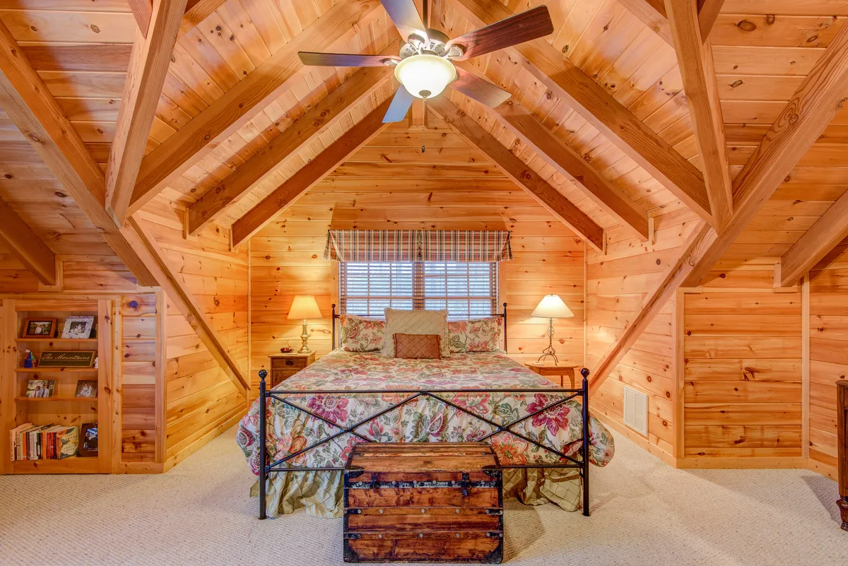 This image shows a bedroom with floral bedding, wooden beams overhead, and nightstands with lamps on either side of the bed. A trunk sits at the foot of the bed, and the entire space feels warm and inviting, with soft lighting that accentuates the textures of the bedding and natural wood elements.