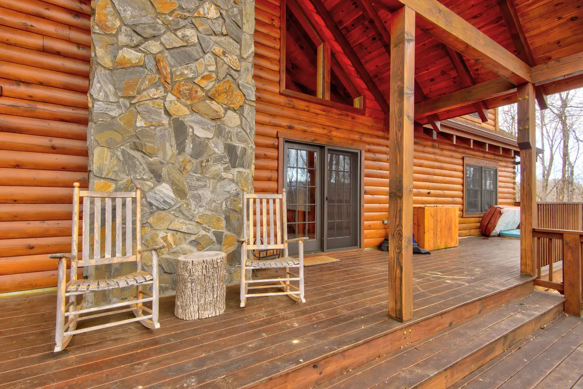 The exterior porch features cozy wooden rocking chairs, large windows, and stone columns, all set against a backdrop of trees. The setting suggests comfort and connection, with spaces ideal for relaxed conversation or quiet moments together.
